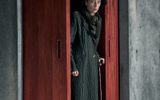 Lia Williams stands in a red doorway with a cane, she wears a long coat and a hat, her face looks wary.