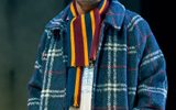 Sebastian De Souza stands with his hands in his coat pockets smiling widely. He wears a patterened winter coat, a beanie hat and a stripey scarf.