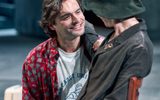 Sebastian De Souza kneels and smiles up at Lia Williams who sits on a chair. He wears a red patterned shirt and she a long ocat and hat, her hand rests on his shoulder warmly.