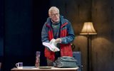 Michael Simkins stands holding a stack of papers, he wears a red and navy coat and a warm jumper with glasses.