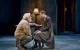 Simon Russell Beale and Lia Williams, Lia sits on a wooden chair and Simon crouches in front of her, they look at each other intently.