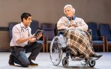 A low shot, in the blurred background are hospital chairs lined against the back wall. In the middle of the stage is Simon Williams in a wheelchair with a tartan blanket over his legs, he has headphones round his neck. He's looking at Sacha Dhawan who is on his knee next to him and reading a book to him.