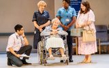 Production photo from Allelujah! The scene visibly a hospital with beige walls and blue signs listing different departments plus faded old chairs. In the middle of the stage is Jacqueline Clarke in a wheelchair. Crouched on the left is Sacha Dhawan in a suit with a stethoscope around his neck. Behind the wheelchair is Deborah Findlay in a dark blue medical smock dress, and next to her is Nicola Hughes in a light blue nurse shirt and dark scrub trousers. On the right is Rosie Ede with neat brown hair and a pink floral mid length dress, she is smiling at Jacqueline.