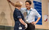 Close shot against the back wall of the set with the blue hospital signage. We see the back of Deborah Findlay in her blue nurses smock. With his arm outstretched leaning on the wall is Gary Wood in a shirt and suit trousers, he has a hospital lanyard on. He's smiling cheekily at Deborah.