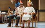 In the background is the remains of the party, there's a balloon with the number 80 on and an older lady eating cake. In the foreground Louis Mahoney (in a striped shirt and jeans but with a crutch in an orange party hat) and Cleo Sylvestre (in a hospital gown and a yellow party hat). Cleo is smiling and Louis is looking at her.