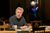 Sthbry-002 Alex Jennings sits on a wooden chair at a wooden table. He wears a grey jumper and holds a pair of glasses