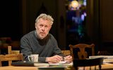 Alex Jennings sits on a wooden chair at a wooden table. He wears a grey jumper and holds a pair of glasses