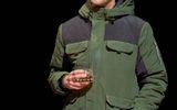 Josh Finan stands behind wooden table, he wears a green coat and holds a glass in one hand and the other in his pocket. His mouth is slightly open and looks affronted.