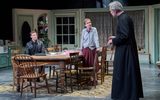 Phoebe Nicholls stands and leans on a table talking to Alex Jennings wearing a black robe. She wears a grey cardigan and long skirt. Jack Greenlees sits at the table watching the exchange.