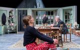 Jo Herbert sits on an ottoman looking up, in the background Alex Jennings and Jack Greenlees can be seen.