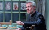 Alex Jennings sits at a wooden table wearing a black blazer, hiw wedding band can be seen on his finger as he clenches his fist whilst speaking.