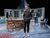 Sthbry-130 Sarah Twomey stands in front of a large wooden table, she wears a puffer jacket, Josh Finan stands behind her out of focus, watching her.