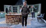 Sarah Twomey stands in front of a large wooden table, she wears a puffer jacket, Josh Finan stands behind her out of focus, watching her.